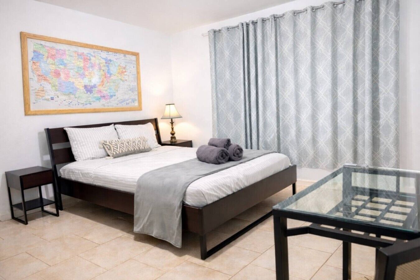 A neatly arranged bedroom with a bed, bedside tables, a glass table, a map on the wall, and a window with curtains.
