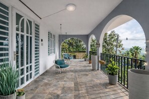 Un espacio exterior techado con suelo de baldosas, ventanas de marco blanco y vistas al verdor.
