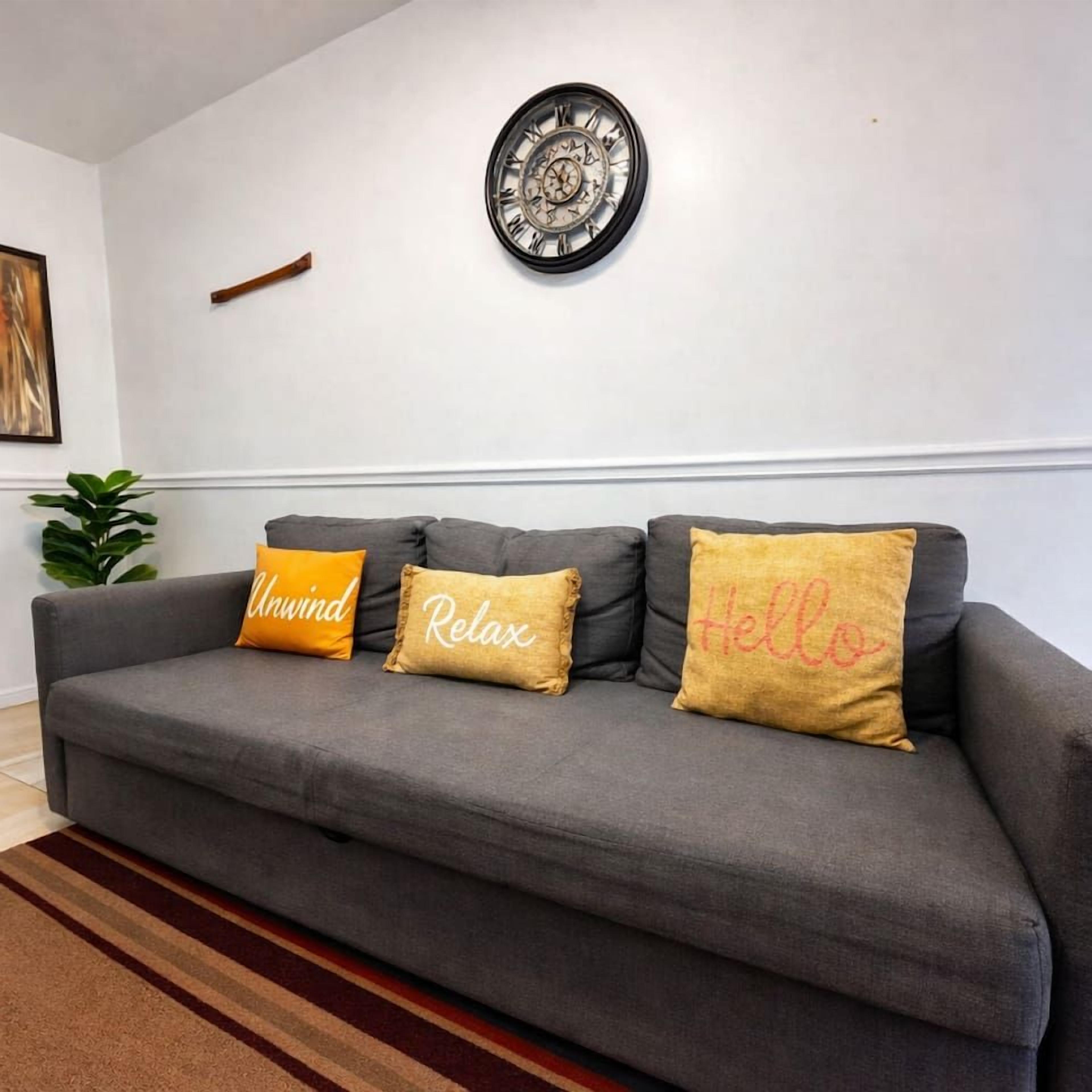 A grey sofa with three cushions, a wall clock, and a plant.