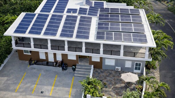 Un immeuble à plusieurs étages doté de panneaux solaires sur le toit, d’un espace de stationnement et d’un petit jardin avec des plantes.
