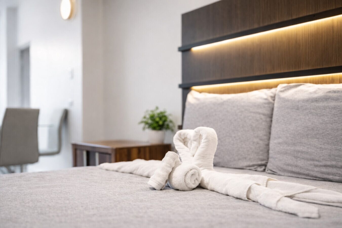 A neatly made bed with a rolled towel, a wooden headboard, and a small table with a plant.