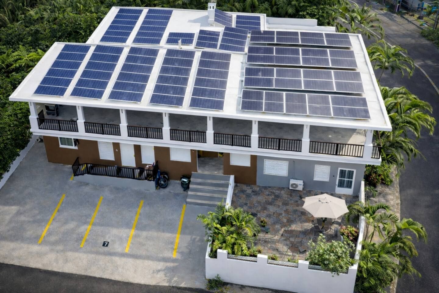 A multi-story building with solar panels on the roof, a parking area, and a small garden with plants.