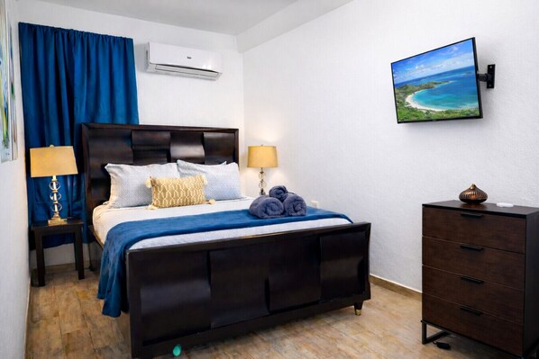 A bedroom with a bed, bedside lamps, a dresser, an air conditioning unit, and a wall-mounted TV. - Nature Glow Access | Pets OK, Parking, AC & WiFi (Ceiba)