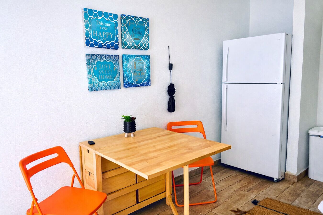 A small kitchen with a wooden table, two orange chairs, a white refrigerator, and four blue wall art pieces.