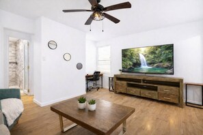Uma sala de estar com uma mesa de centro de madeira, uma televisão sobre um suporte de madeira, um ventilador de teto e uma parede de pedra.