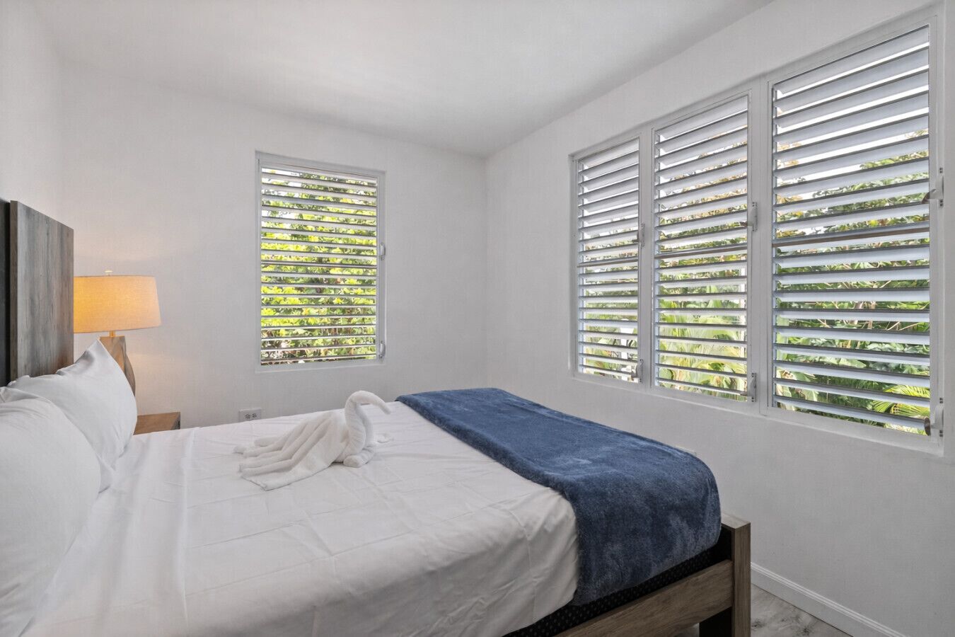 A bedroom with a bed, two windows with blinds, a lamp, and a wooden headboard.