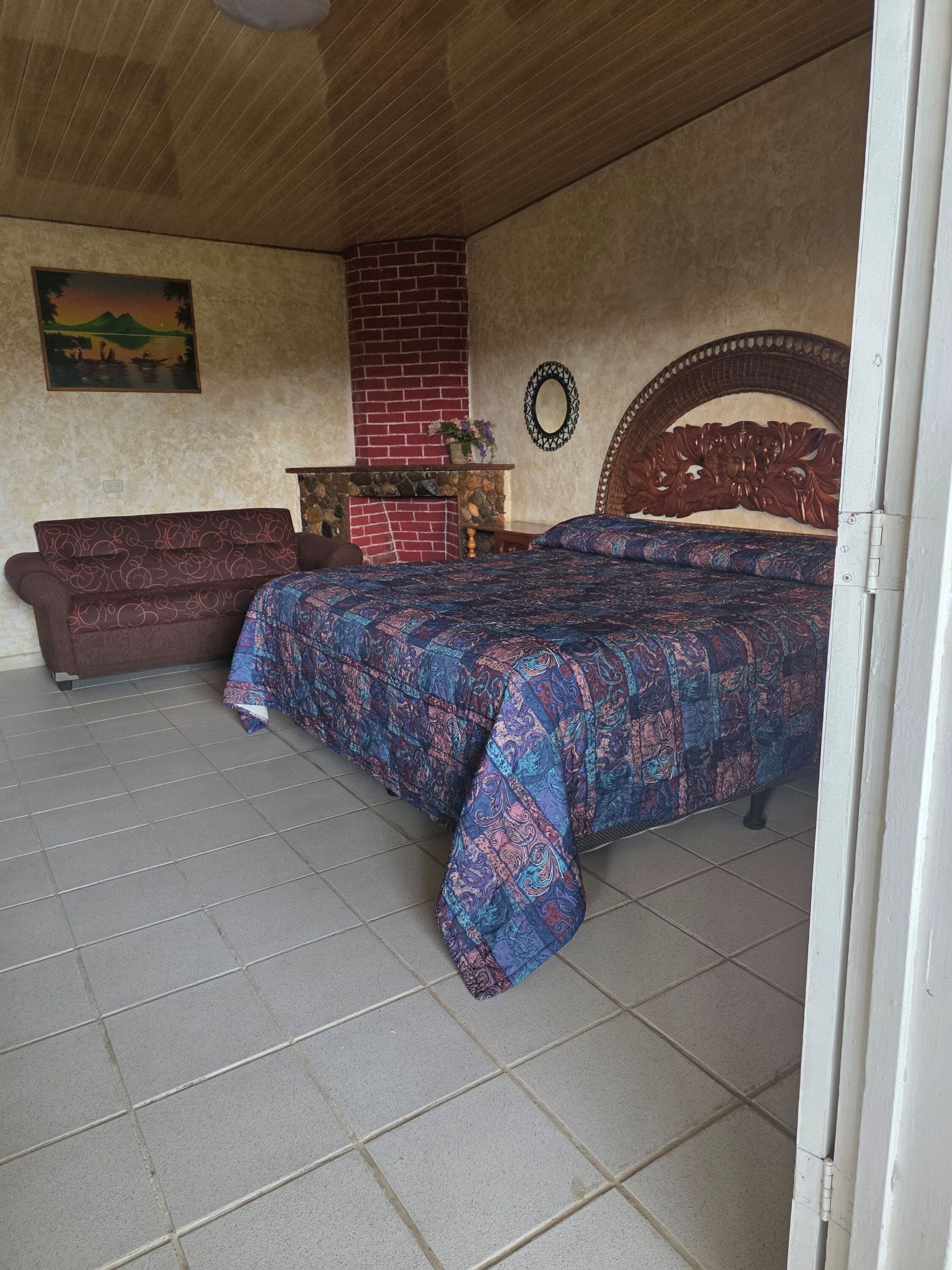A bedroom with a bed, a wooden headboard, a red brick fireplace, a wooden bench, and a wall-mounted mirror.