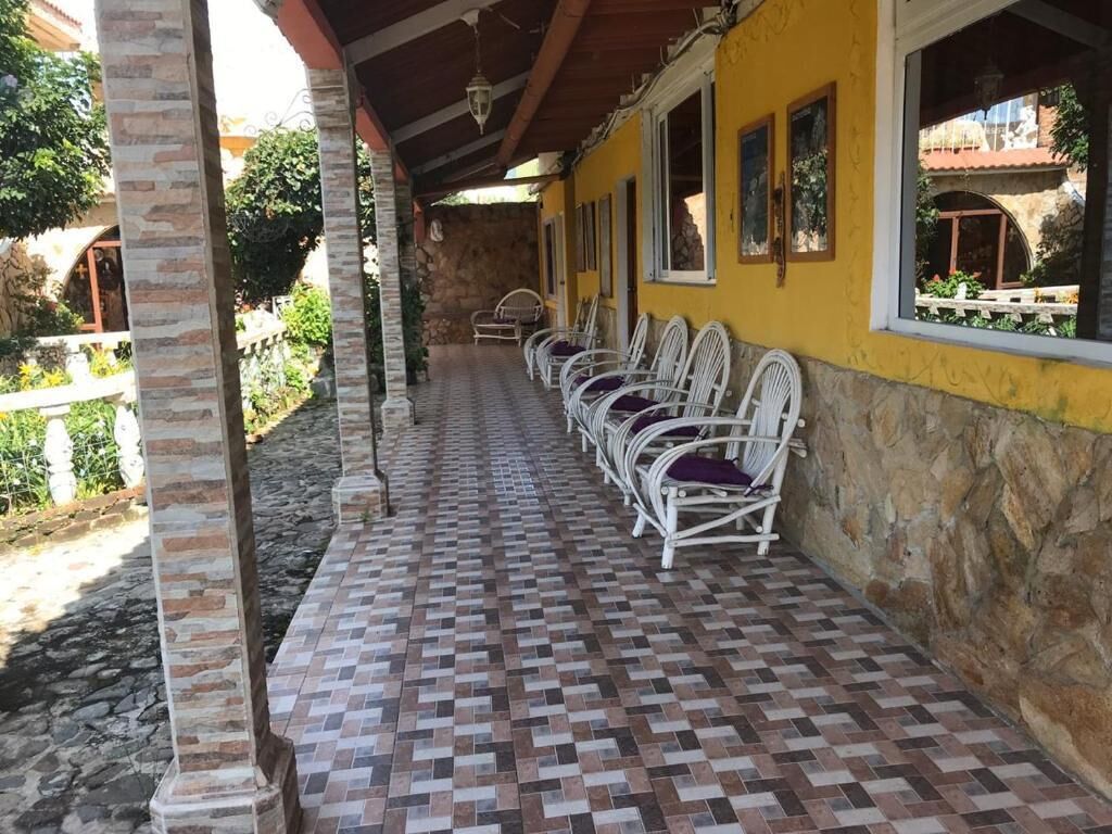 A covered outdoor seating area with a tiled floor, white chairs with purple cushions, and a yellow wall with windows.