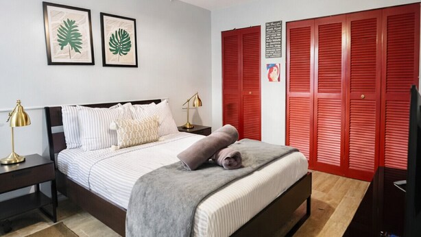 A bedroom with a bed, bedside tables, a desk, a chair, a lamp, a red closet, and a wall-mounted artwork. - Solar Island Base | WiFi, Parking and Near Ferry (Ceiba)