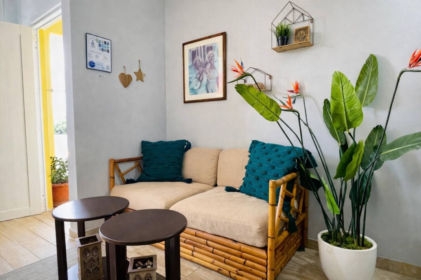 A cozy room with a sofa, two small tables, a large potted plant, and wall decorations including a framed picture and a framed certificate.