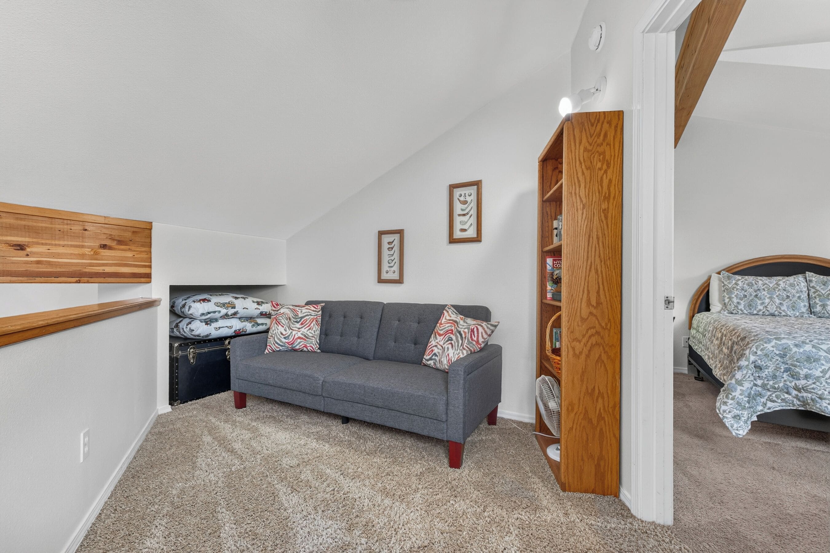 Ein Schlafzimmer mit grauem Sofa, einer Holzbücherregal, einem Bett mit gemusterten Betttextilien und einem kleinen Tisch mit einer Lampe.