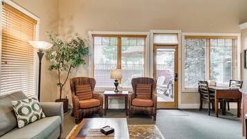 Un salon comprenant un canapé, deux fauteuils, une table basse et une lampe. De grandes fenêtres équipées de stores laissent entrer la lumière, et on aperçoit une plante ainsi qu’une vue sur l’extérieur.