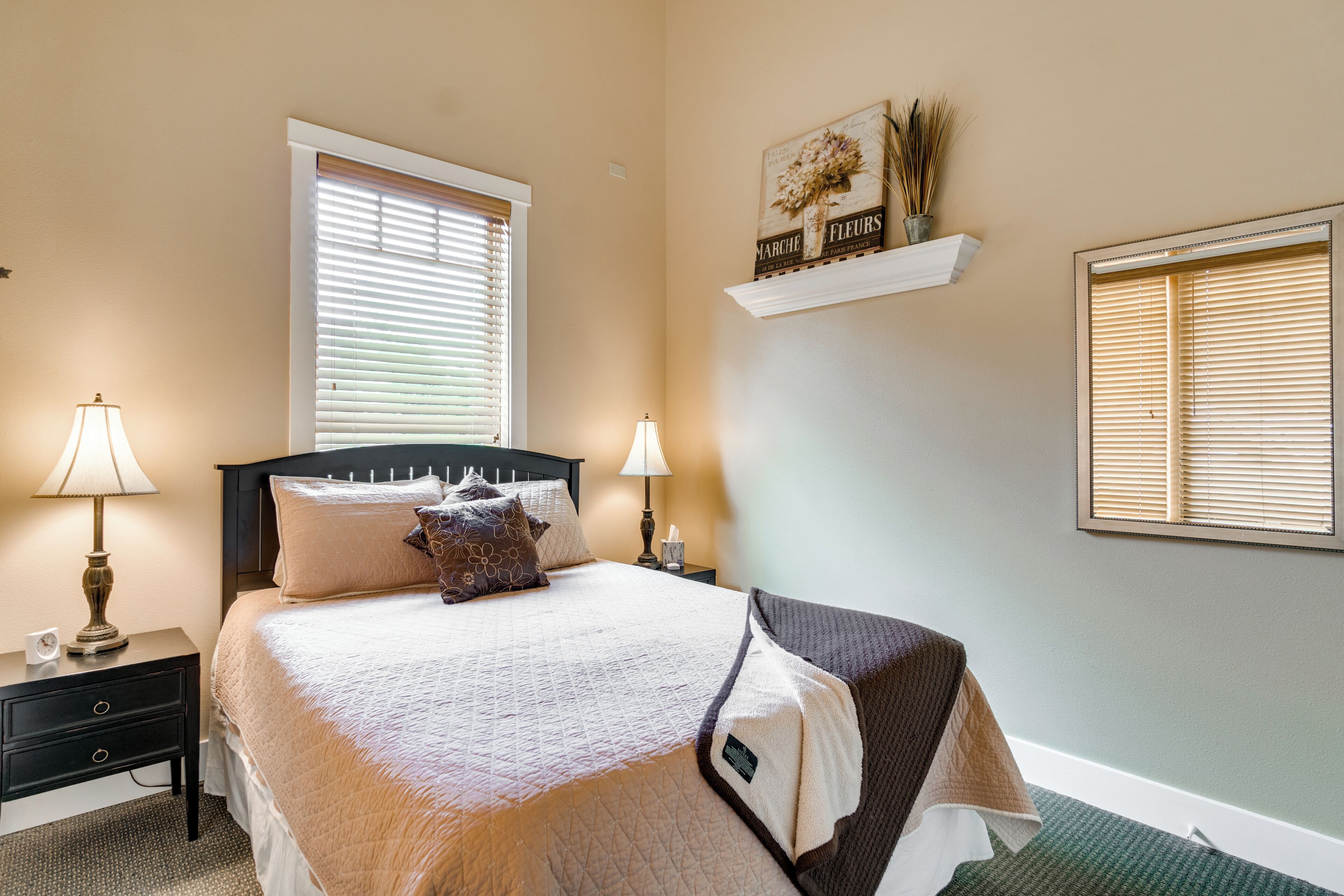 Une chambre à coucher bien rangée, avec un lit, deux tables de chevet dotées de lampes, une table de nuit, une fenêtre avec des stores et une étagère murale agrémentée d’objets décoratifs.