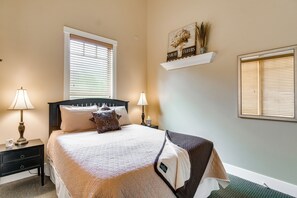 Un dormitorio ordenado con una cama, dos mesitas de noche con lámparas, una mesita auxiliar, una ventana con persianas y un estante de pared con objetos decorativos.
