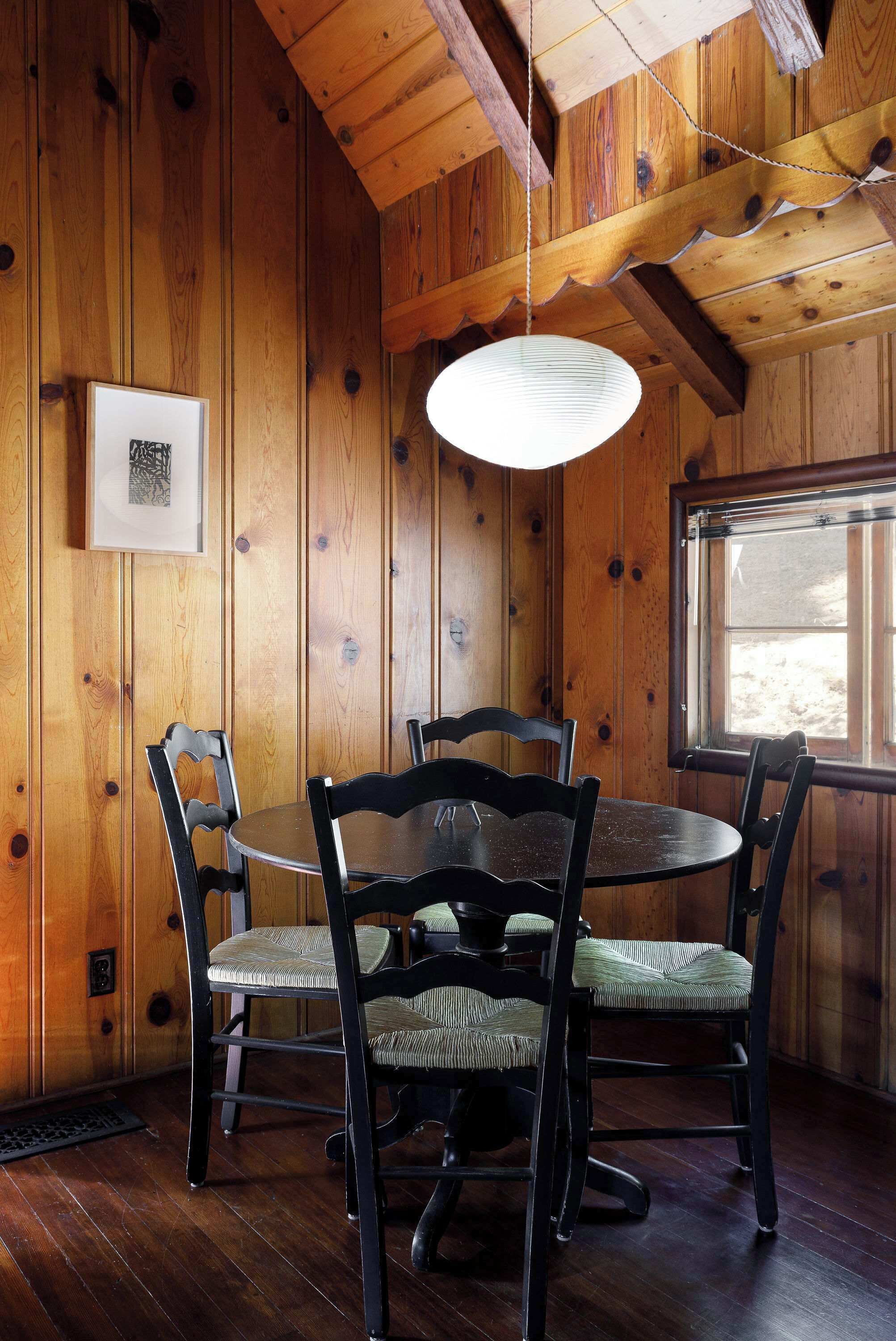 A wooden interior with a round table and chairs, a hanging light fixture, and a window.