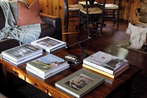 Ein Holztisch mit mehreren Büchern, eine schwarze Stehlampe und eine gemütliche Wohnzimmergestaltung mit Sofa und Sesseln.