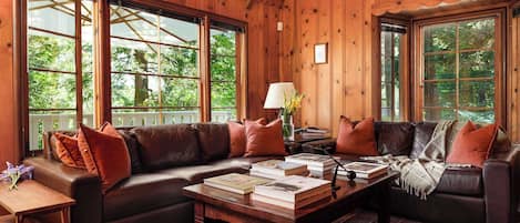 A wooden interior with a sectional sofa, a coffee table with books, and large windows.