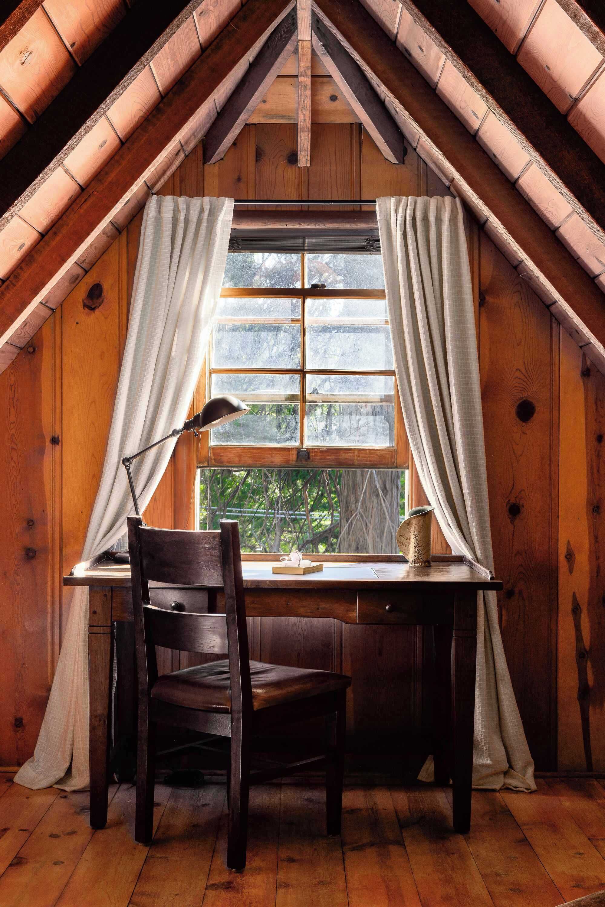 A wooden room with a sloped ceiling, a desk with a chair, and a window with curtains.