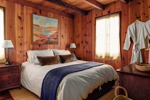 A wooden-paneled bedroom with a bed, two bedside lamps, a painting, and a robe hanging on the wall. - The Writer's Cabin in Lake Arrowhead Woods (Lake Arrowhead)