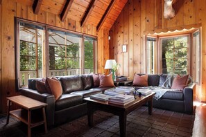 Ein gemütliches Cabin-Interieur mit einer Ledersofa, einem Couchtisch mit Büchern und großen Fenstern mit Blick auf den Wald.