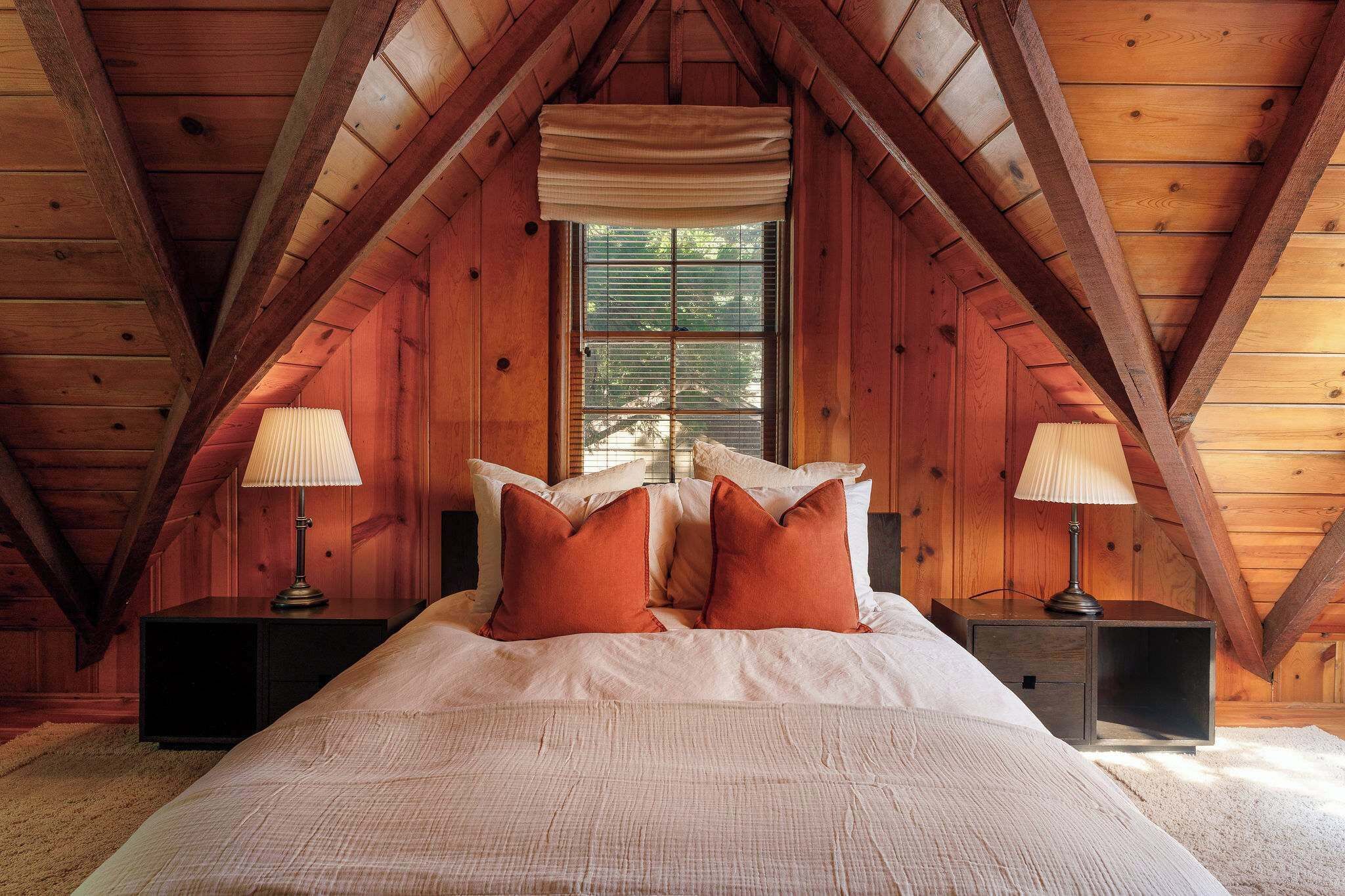 A bedroom with a sloped ceiling, a large bed with pillows, two bedside tables with lamps, and a window with blinds.