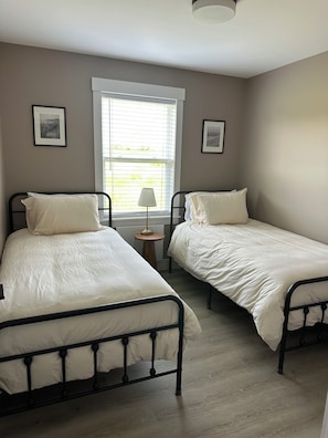 Een slaapkamer met twee enkele bedden, een raam met zonwering, een kleine tafel met een lamp en lijstjes met foto’s aan de muur.