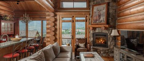 A log cabin interior with a stone fireplace, a sofa, a dining area, and a view of the mountains through the windows.