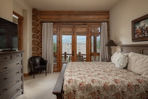 Ein Schlafzimmer mit Bett, Kommode, Sessel, Fernseher und Blick auf Berge durch große Fenster.