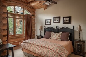 A log cabin-style bedroom with a large window, a bed with floral bedding, two bedside lamps, and three framed pictures on the wall. - Luxury Mountain Village Retreat | Panoramic Mountain Views | Private Hot Tub | Sleeps 10 (Telluride)