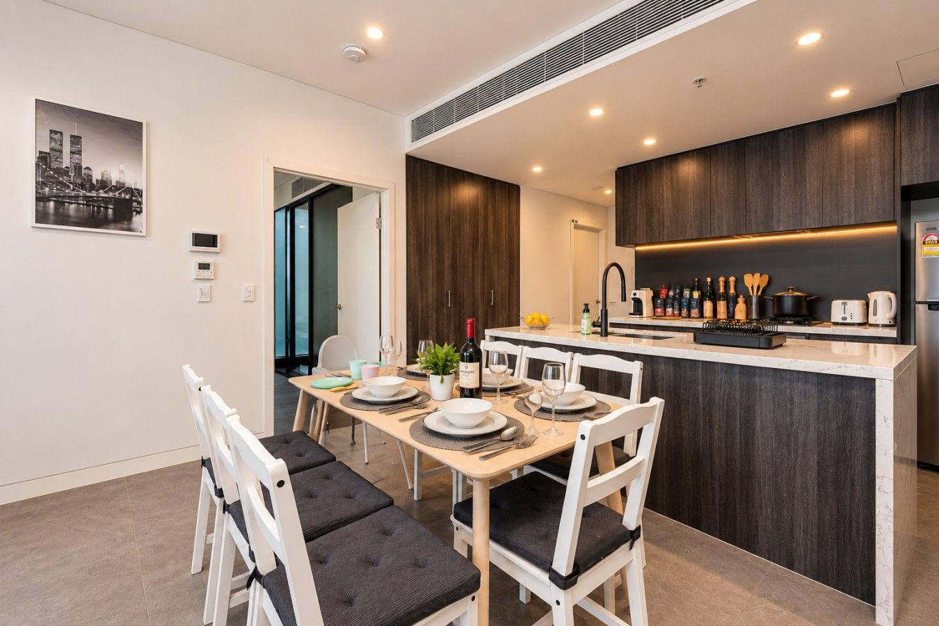 A modern kitchen with dark wood cabinetry, a central island, and a dining area with white chairs and a table set for two.