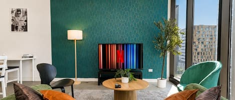 Una sala de estar moderna con una pared estampada en color turquesa, un televisor de pantalla plana, una mesa de centro redonda y un sofá verde con cojines naranja.