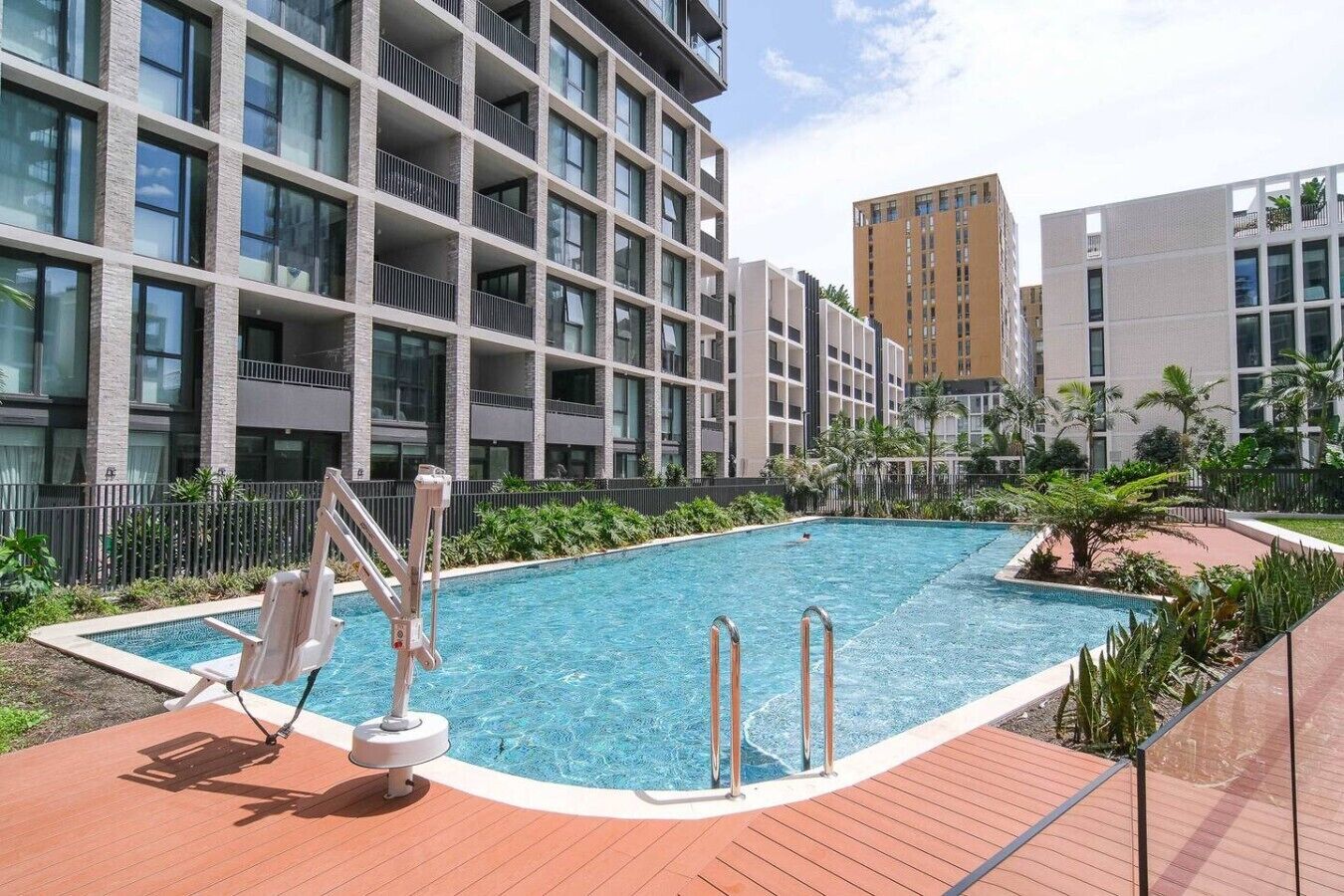 A swimming pool with a wooden deck, surrounded by a fence, and adjacent to a multi-story building with balconies.