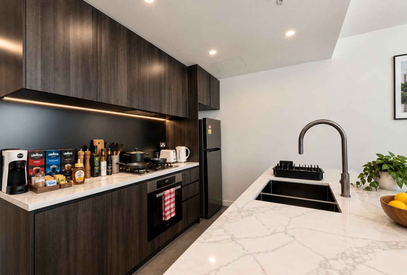 Kitchen corner showing the four burner gas cooktop on the bench against the wall with the oven below it, dark cabinetry with a marble look benchtop, and under cabinet lighting. The island sink and its gooseneck tap are in the foreground.
