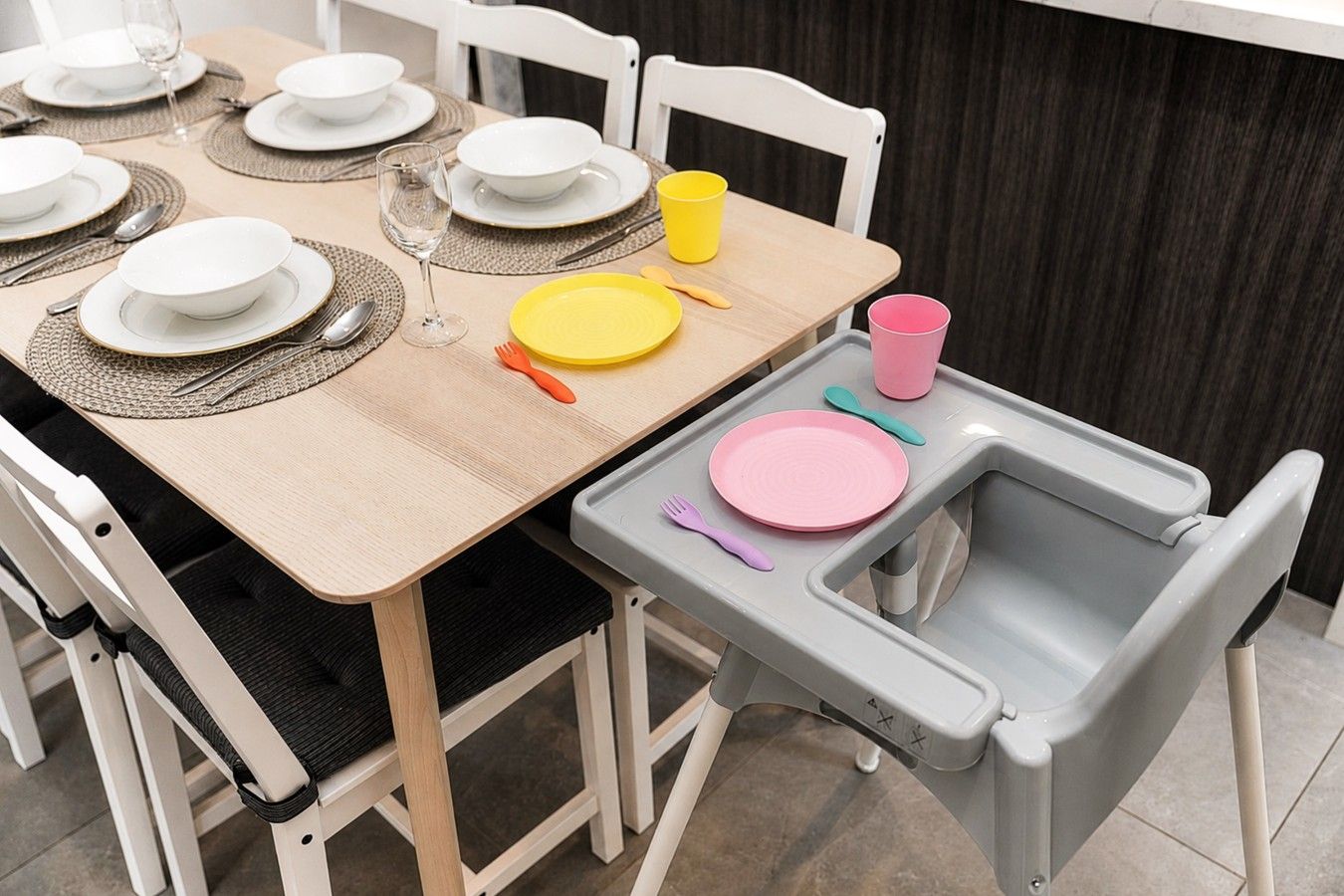 Una mesa de comedor dispuesta con platos, cubiertos y vasos, además de una trona infantil con bandeja.