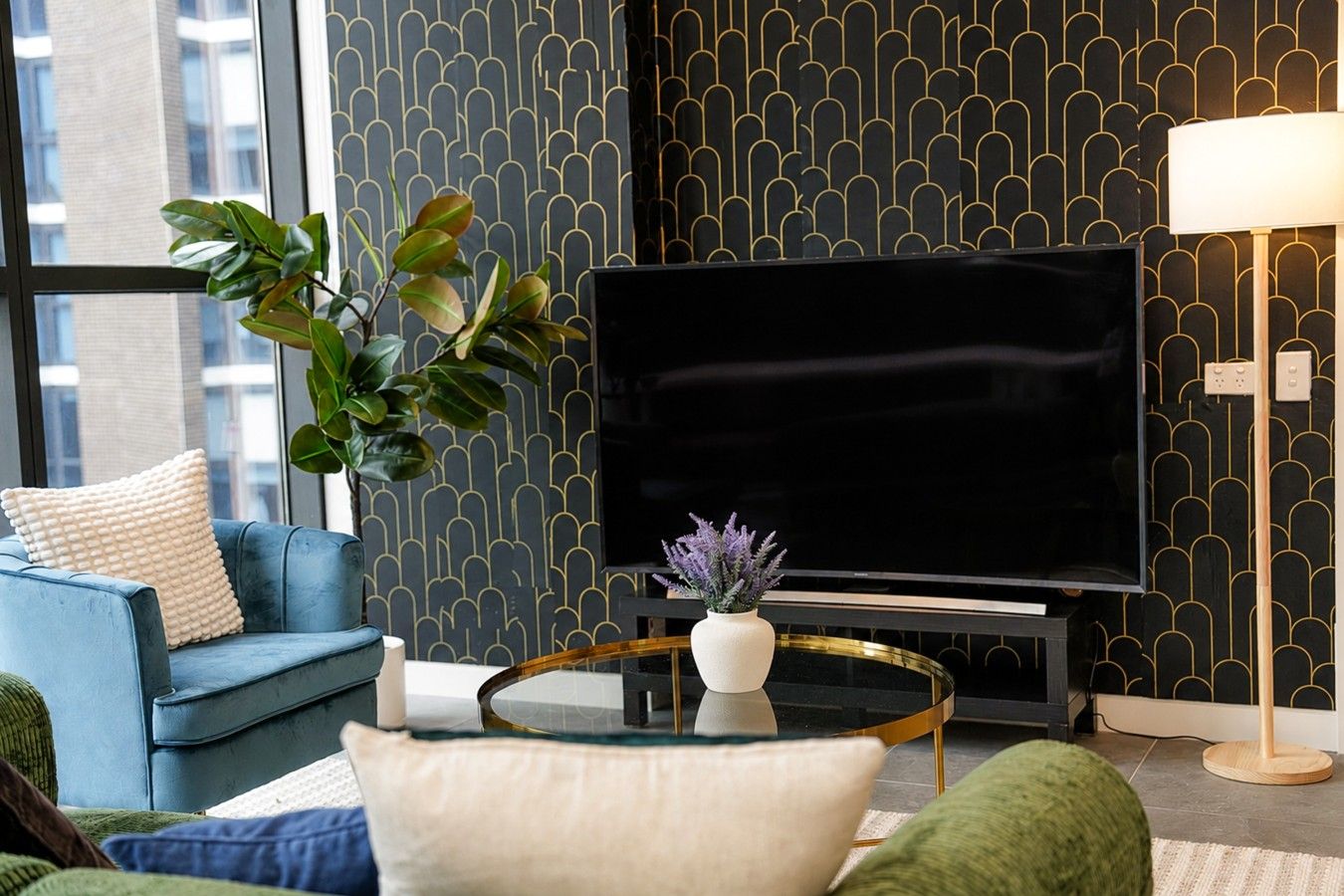 En moderne stue med en blå sofa, et rundt sofabord, et fladskærms-tv, en potteplante og en gulvlampe.