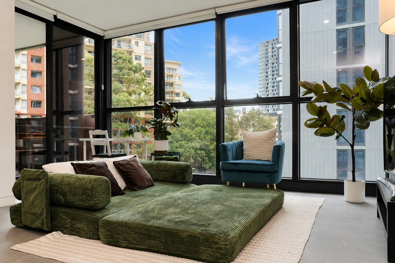 A modern living room with a green sofa set, a blue armchair, and a large window offering a city view.