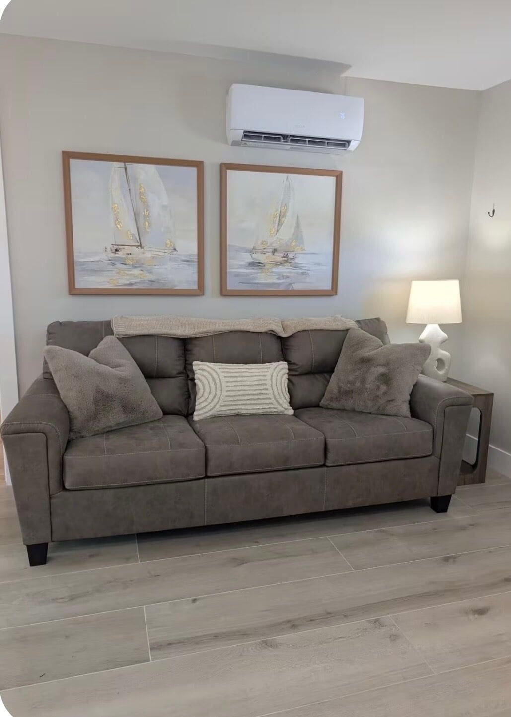 Un salon avec un canapé gris, deux tableaux encadrés, un climatiseur blanc et une lampe.
