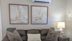 A living room with a grey sofa, two framed paintings, a white air conditioner, and a lamp. - The Eden (Fort Myers Beach)