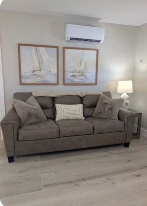 A living room with a grey sofa, two framed paintings, a white air conditioner, and a lamp. - The Eden (Fort Myers Beach)