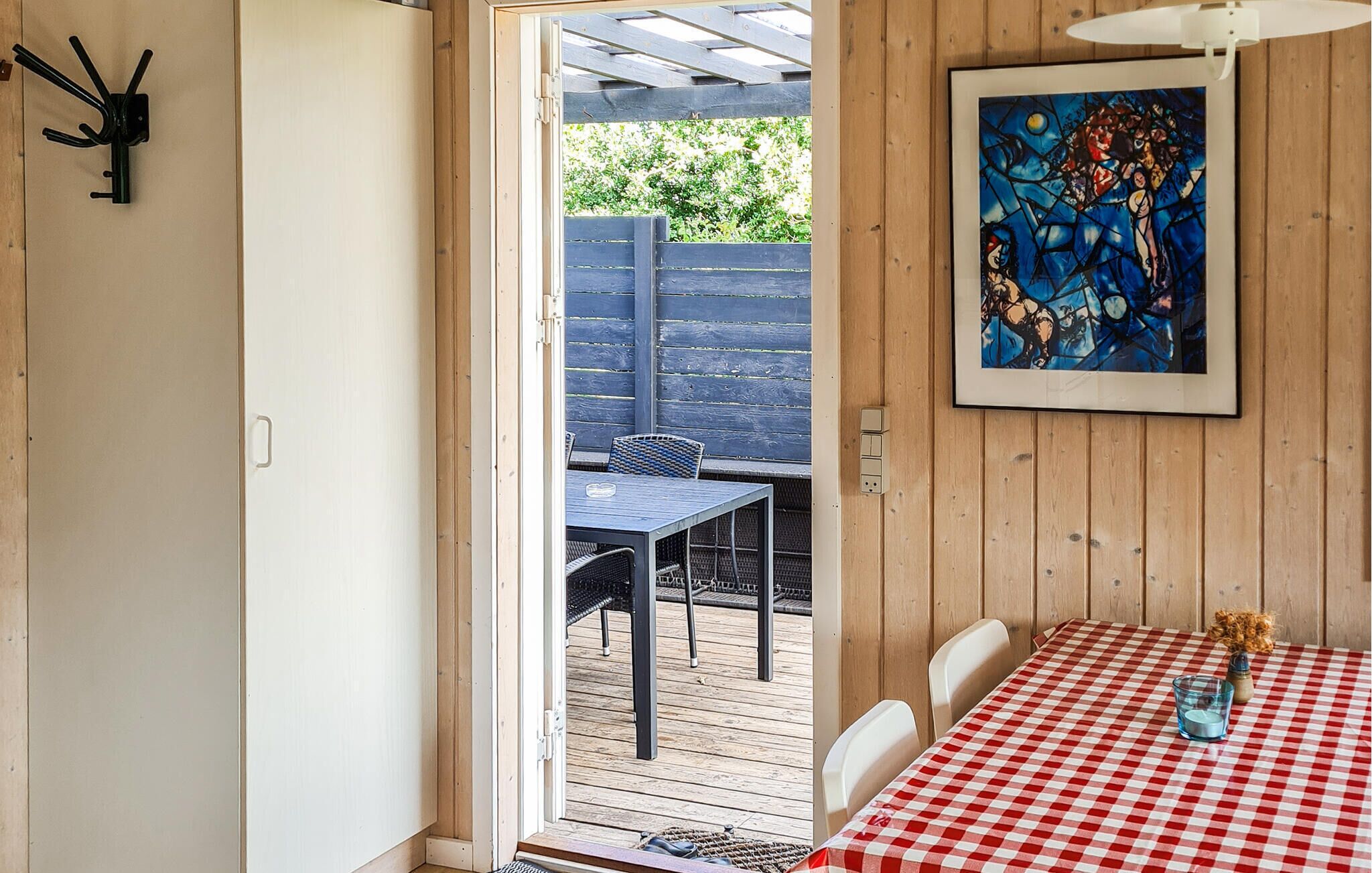 A room with a checkered table, a framed abstract painting, and a view of an outdoor patio with a table and chairs.