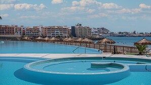 Uma grande piscina com design circular e águas cristalinas, rodeada por um deck de madeira e guarda-sóis de palha. Ao fundo, edifícios altos com varandas e vista para o mar.