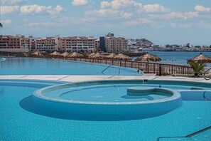 Ein großer, klarer Pool mit kreisförmigem Design, umgeben von einer Holzterrasse und Reetdachschirmen. Im Hintergrund befinden sich mehrstöckige Gebäude mit Balkonen und Blick auf das Meer.