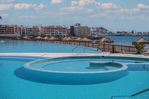 A large, clear swimming pool with a circular design, surrounded by a wooden deck and thatched umbrellas. In the background, there are multi-story buildings with balconies and a view of the sea. - Two 703 : The Coastal Gem (Puerto Peñasco)