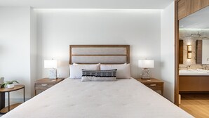Ein ordentlich eingerichtetes Hotelzimmer mit einem großen Bett, zwei Nachttischen mit Lampen, einem kleinen Beistelltisch mit Pflanze und einem durchgehenden Türbogen einsehbaren Badezimmer.