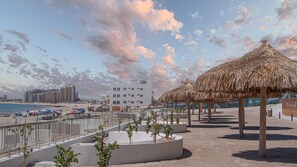 Ein Strandresort mit einem strohgedeckten Pavillon, einem Poolbereich mit Palmen und Blick auf das Meer und die Stadtlandschaft.