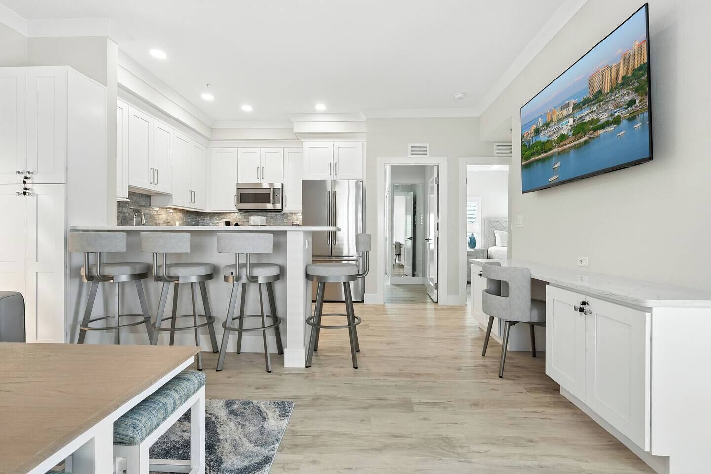 A modern kitchen with white cabinetry, a central island with bar stools, and a large flat-screen TV mounted on the wall.