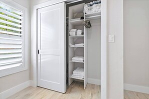 Ein weißer Kleiderschrank mit Regalen und Kleiderstange, ein Fenster mit Jalousien und ein heller Boden.