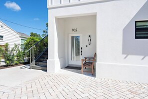 A white building with a number '102' on the door, a wooden bench, and a staircase leading to the entrance.