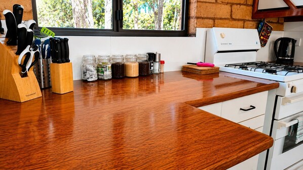 A kitchen with a wooden countertop, a gas stove, a window with a view of trees, and various kitchen utensils and containers. - Yelverton Brook - Sheoak Lodge - 2 bedroom (Metricup)