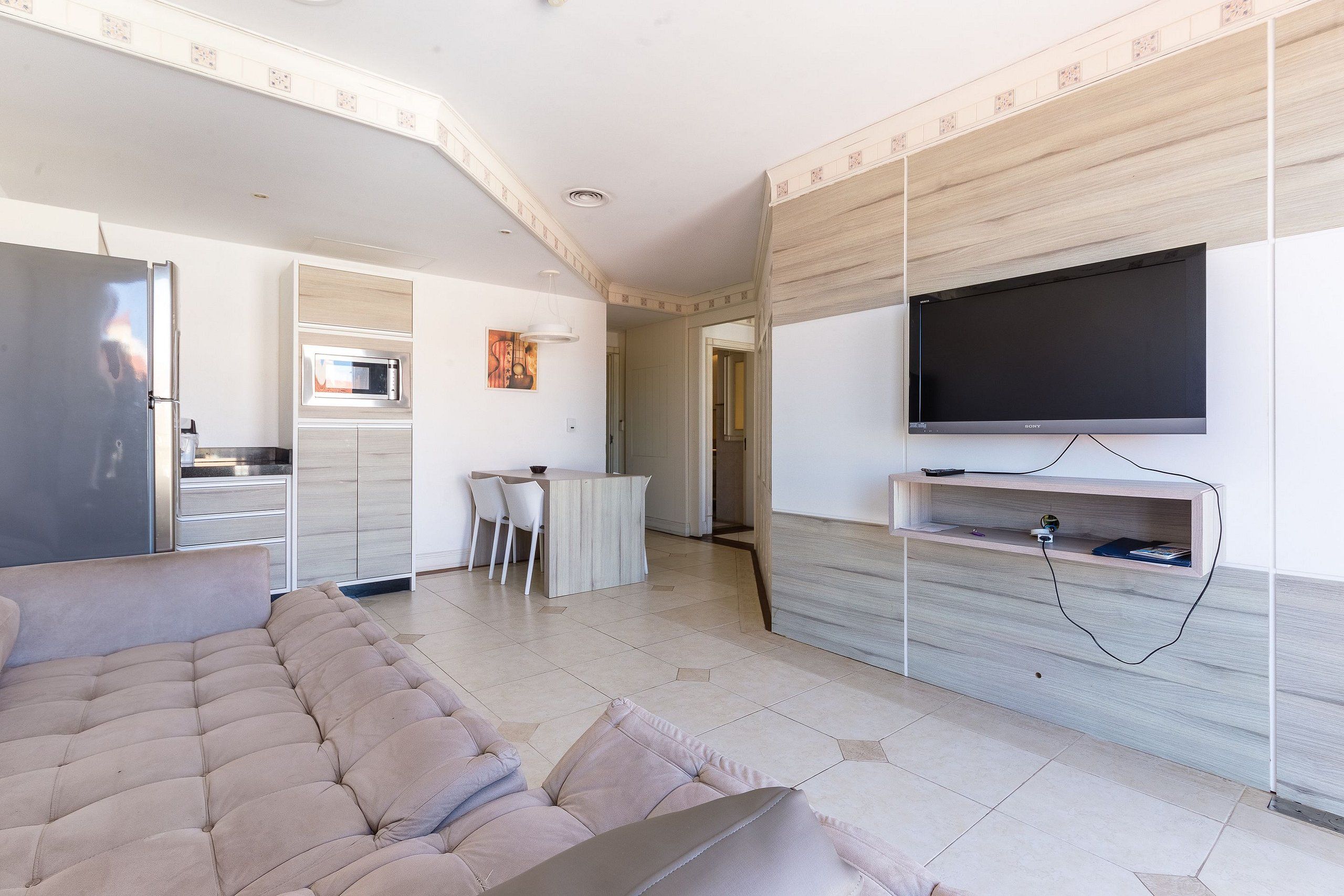 A modern living room with a sofa, a flat-screen TV mounted on a wooden wall, a kitchen area with a microwave, and a dining table with chairs.